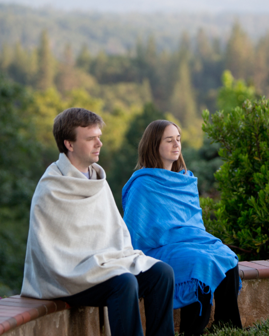 Two people meditating together outside, wearing shawls